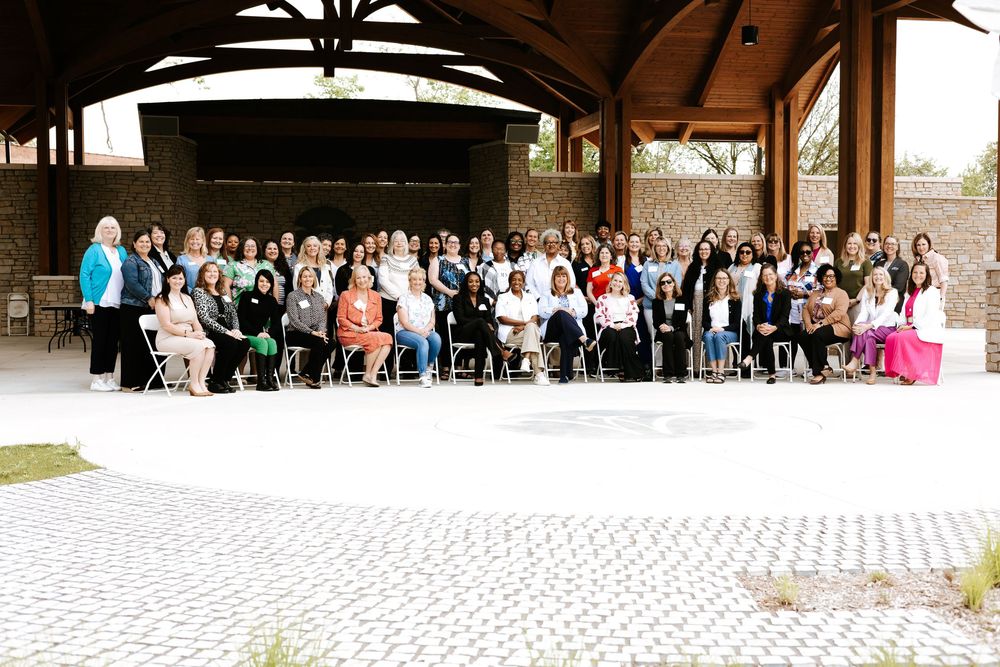 Large group of women posing for a formal group photo under an outdoor pavilion at a professional event.