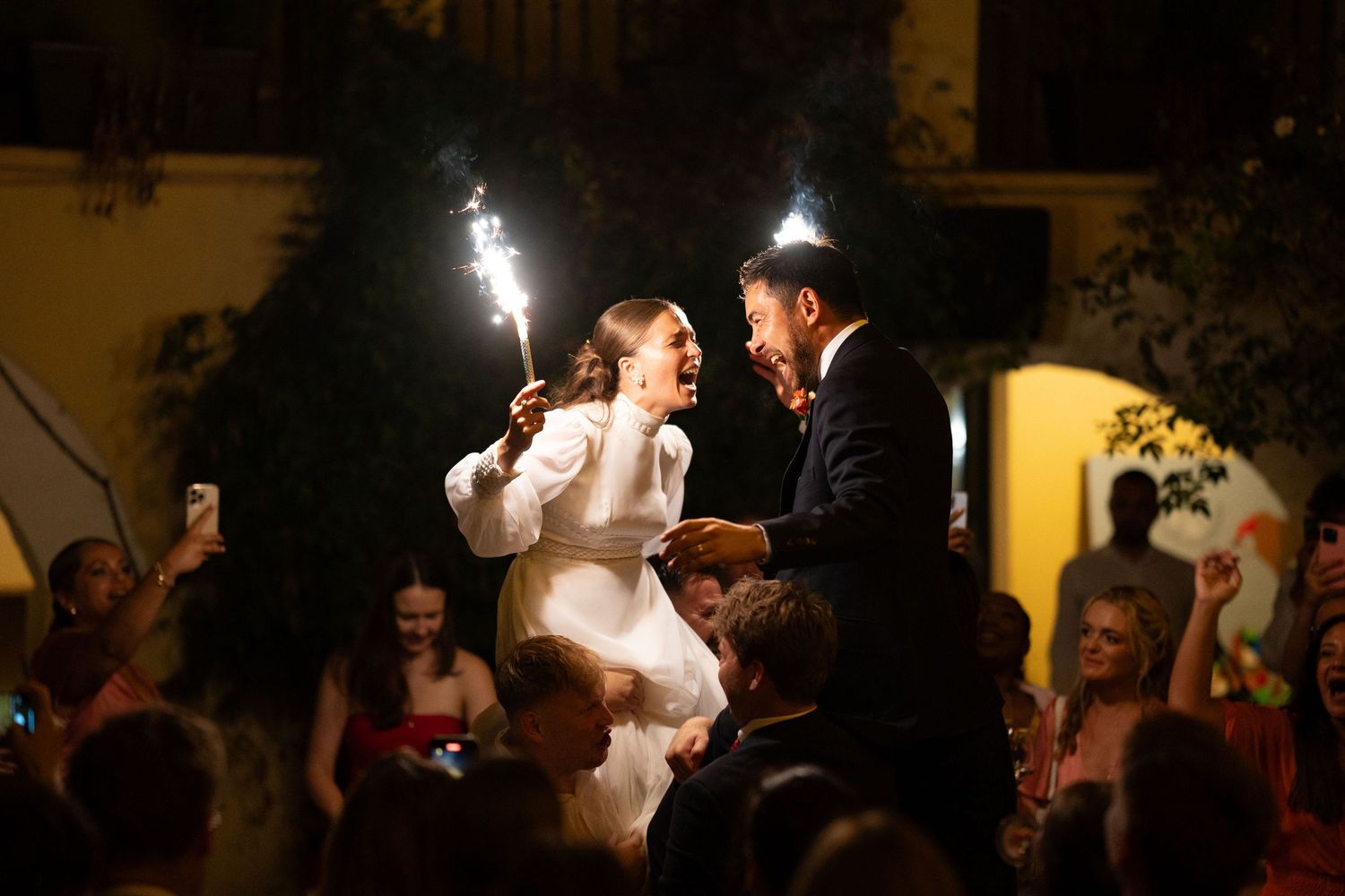 Bride and groom laughing and holding sparklers on the shoulders of their friends and families at night In Sotogrande