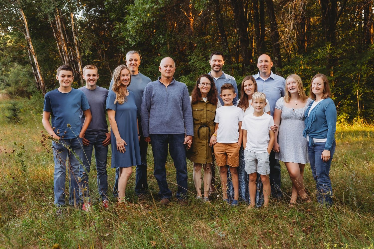 A large family group poses together in a sunny meadow wearing coordinated blue and white outfits.