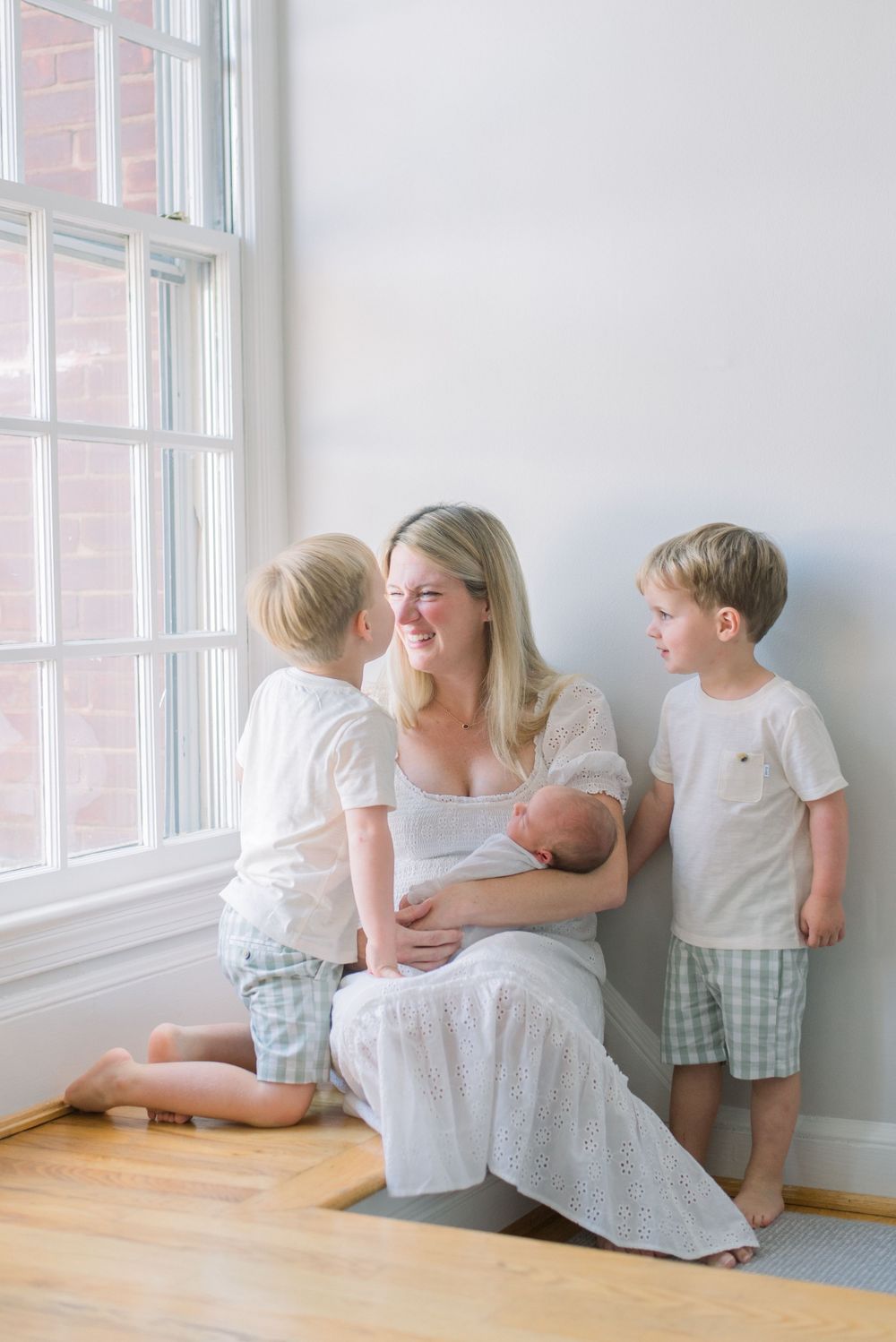 Children sit together by a bright window in a minimalist white room with hardwood floors.