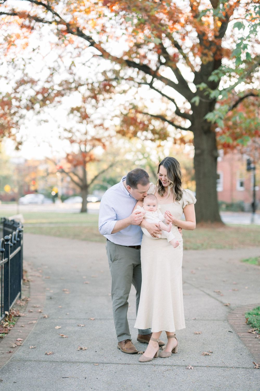 A family shares tender moments together on a walkway surrounded by autumn trees with orange and red foliage.