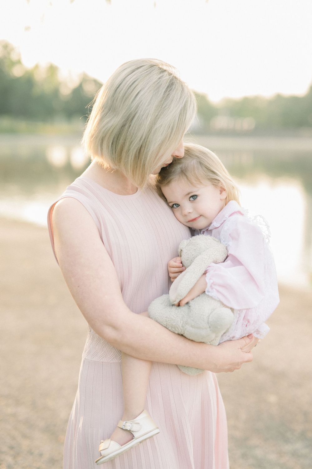 A tender moment captured in soft lighting shows a parent and child embracing in matching pink outfits by a lake at sunset.