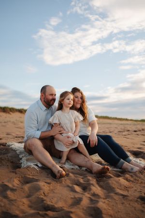 Family sitting together on sandy beach at sunset with cloudy sky in background.