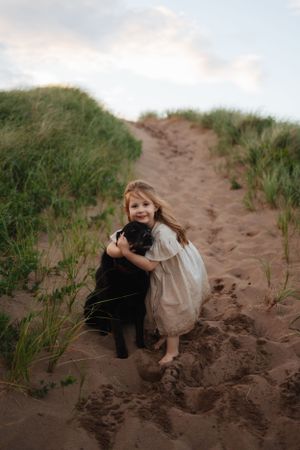 Child in white dress hugging black dog on sandy path through grass-covered dunes.