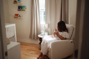 Person in white dress sitting in nursery chair near window with floating shelves.