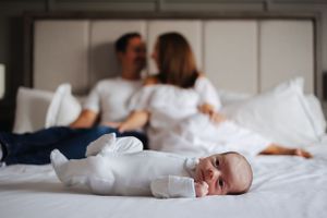 Newborn baby in white onesie lying on bed with parents in background.