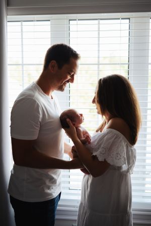 Silhouetted family moment by window with newborn baby in white nursery.