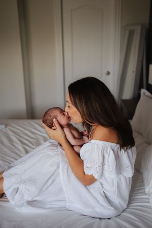 Mother in white dress holding newborn baby tenderly on white bedding.