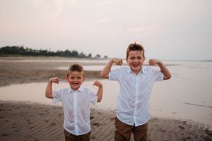 Two children in white shirts pose playfully on a beach at sunset showing off their muscles.