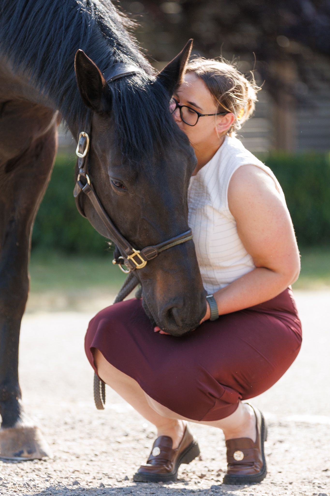 Woman in glasses and a burgundy skirt crouches outdoors, tenderly resting her face against a dark bay horse wearing a leather bridle.