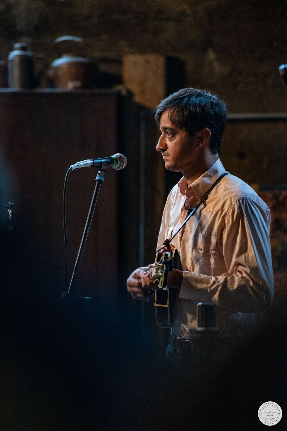 Musician in a light button-up shirt plays an acoustic guitar beside a microphone on a stand during a live performance in a rustic venue.