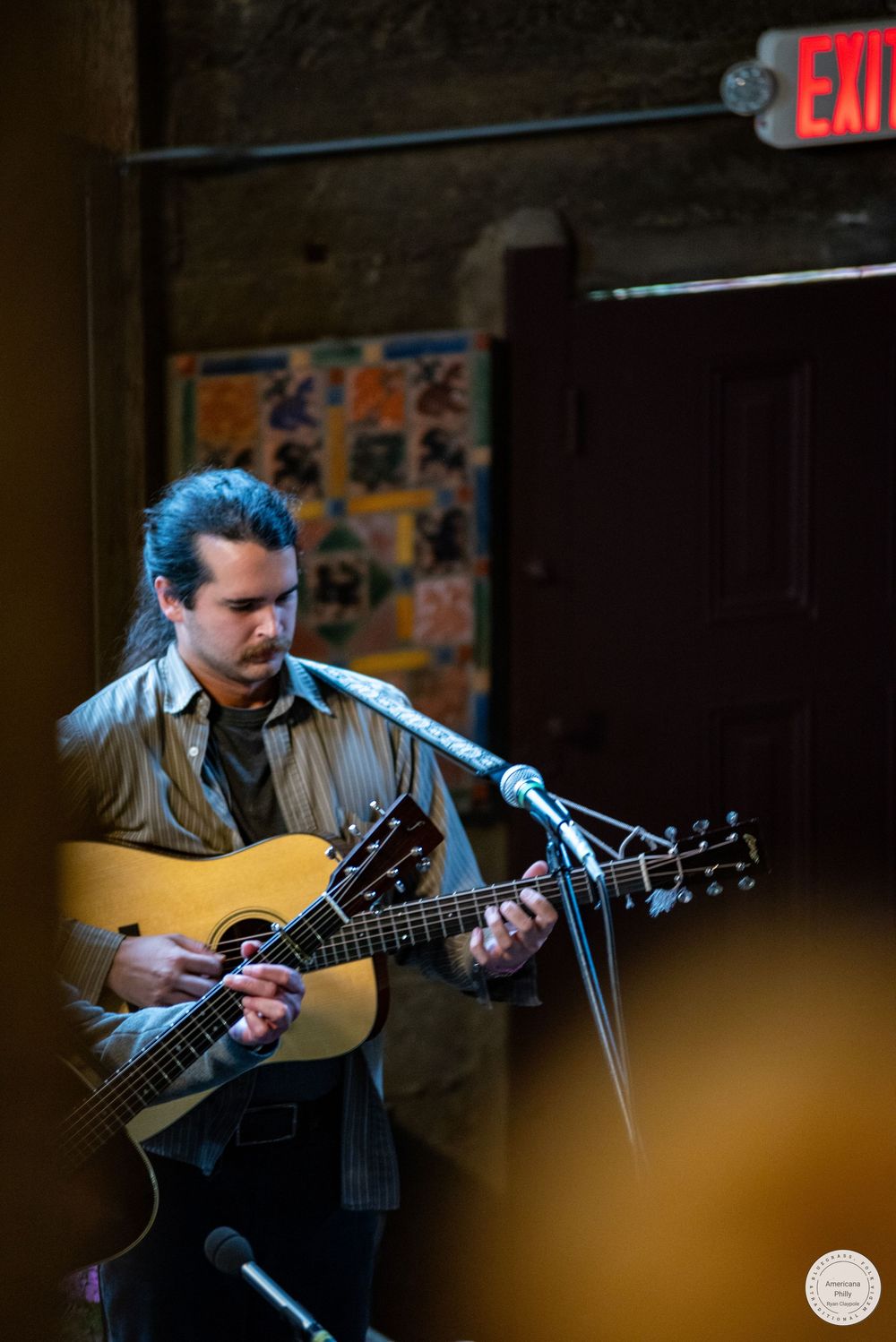 A bearded musician with long dark hair plays an acoustic guitar beside a microphone on a stage in a rustic venue with an exit sign.