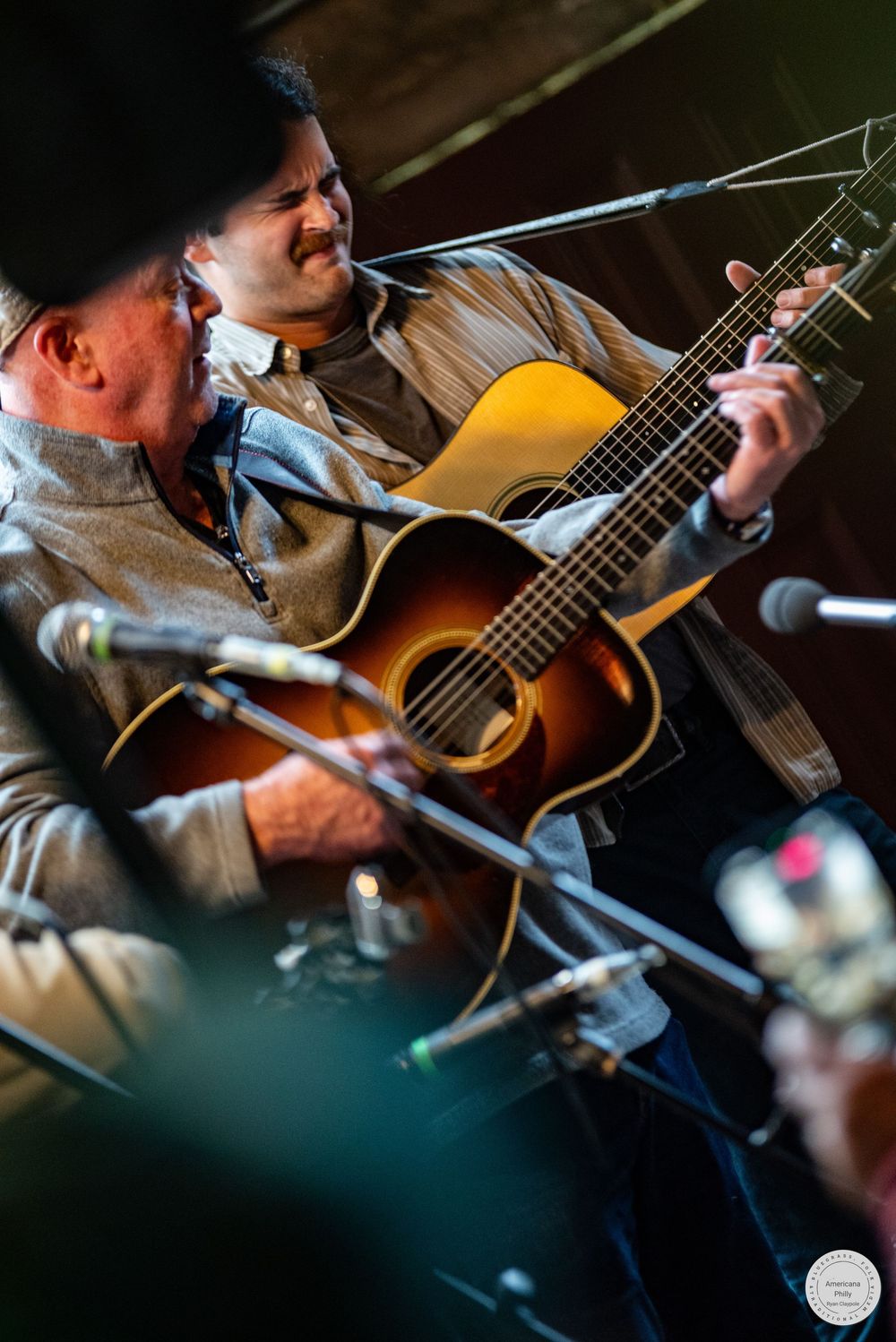 Two musicians play acoustic guitars on stage during a live bluegrass performance, with microphones and stands in the foreground.