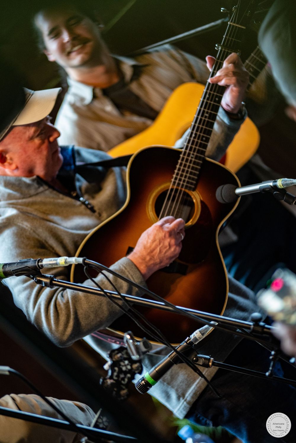Two musicians play acoustic guitars on stage in front of microphones during a live bluegrass or folk performance.