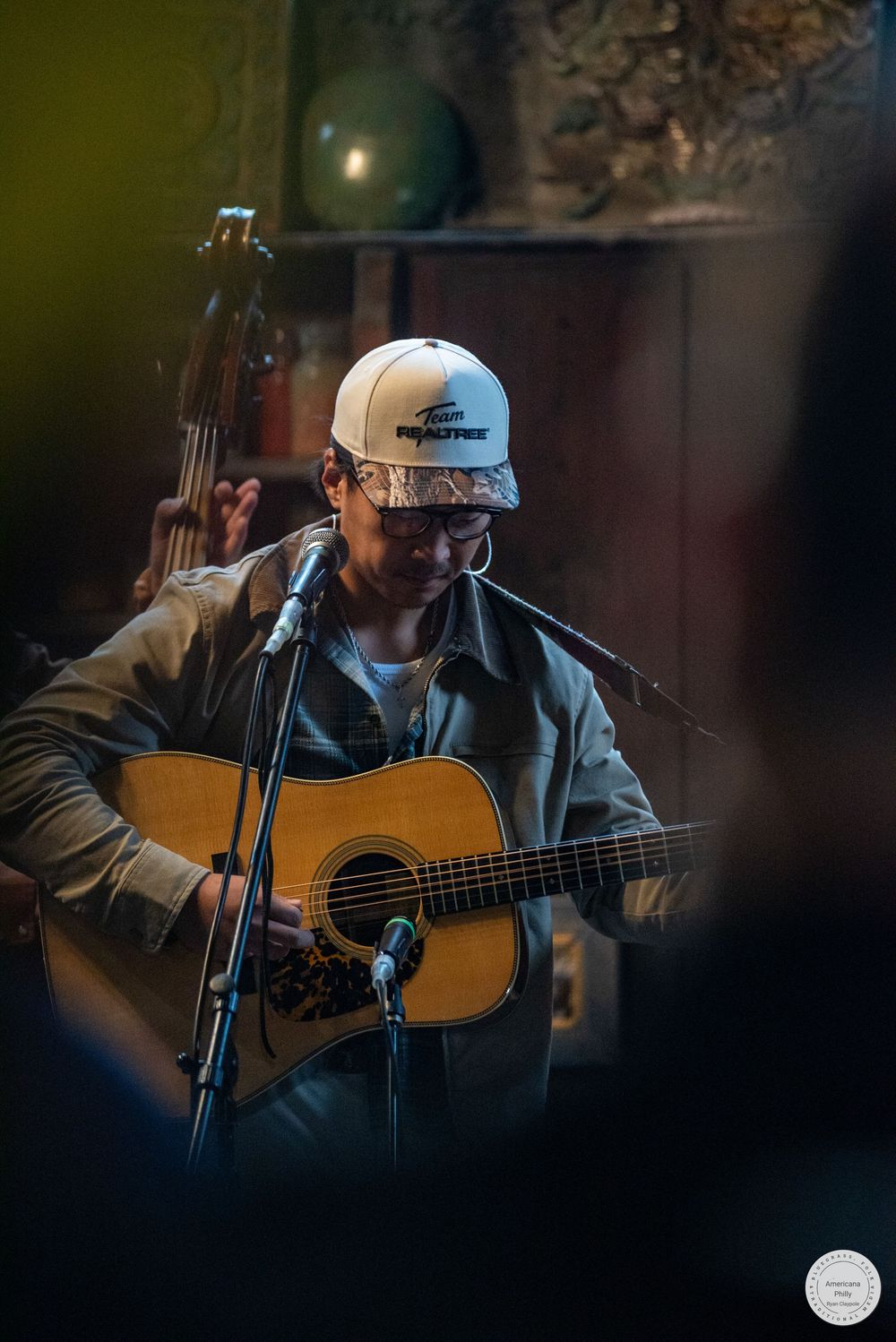Musician in a white cap and glasses plays an acoustic guitar and sings into a microphone during an intimate live performance.