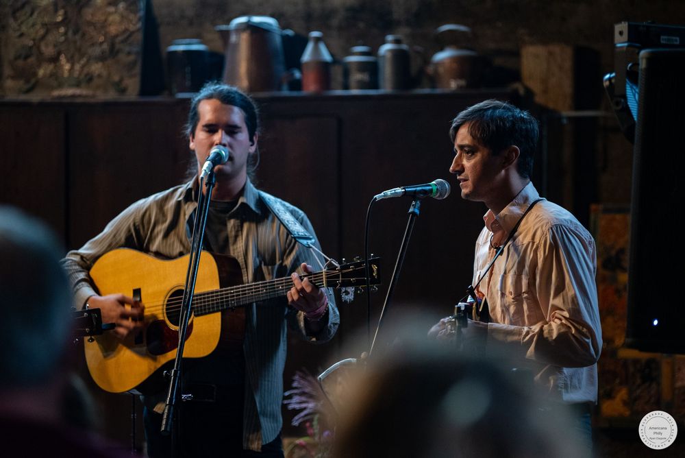 Two musicians perform live on stage, one singing and playing an acoustic guitar at a microphone, with an audience in the foreground.