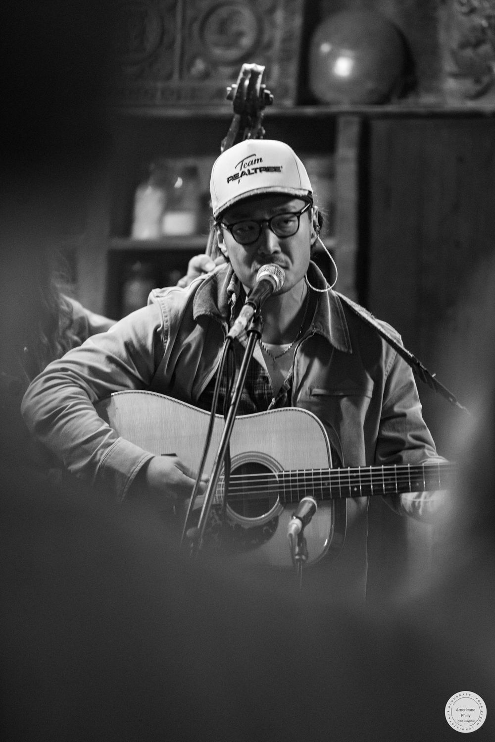 Black and white photo of a musician in a cap and glasses singing into a microphone while playing an acoustic guitar on stage.