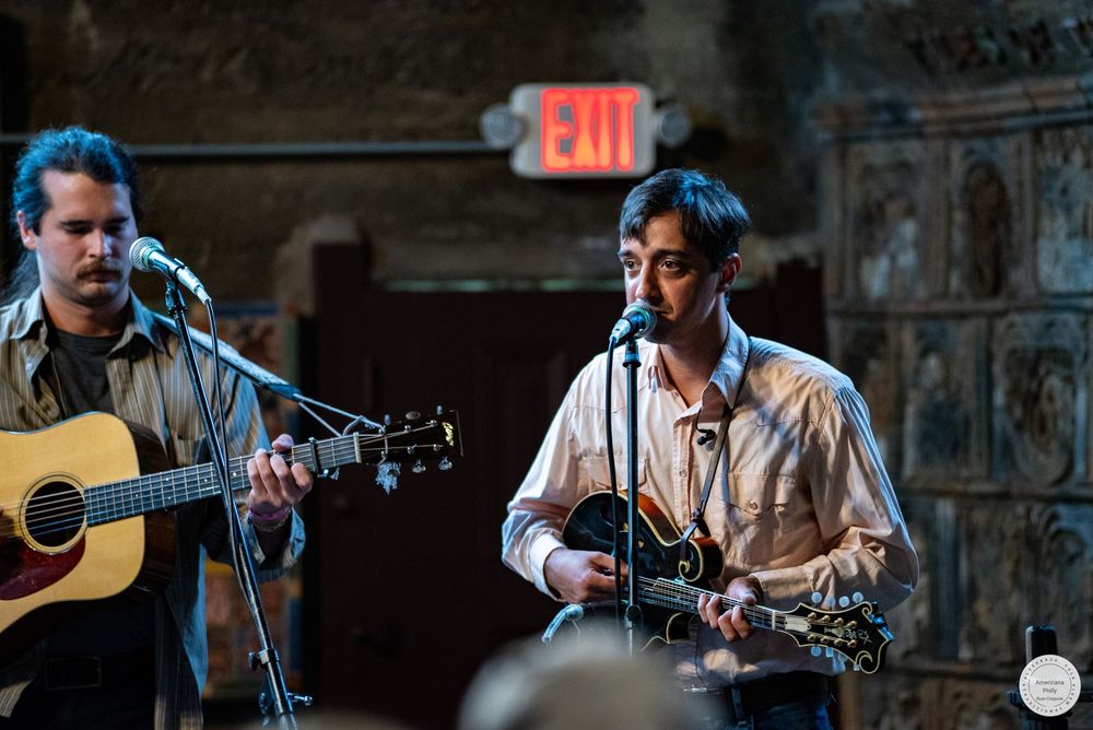Two musicians play acoustic guitars and sing into microphones on a dimly lit stage with an illuminated red exit sign above.