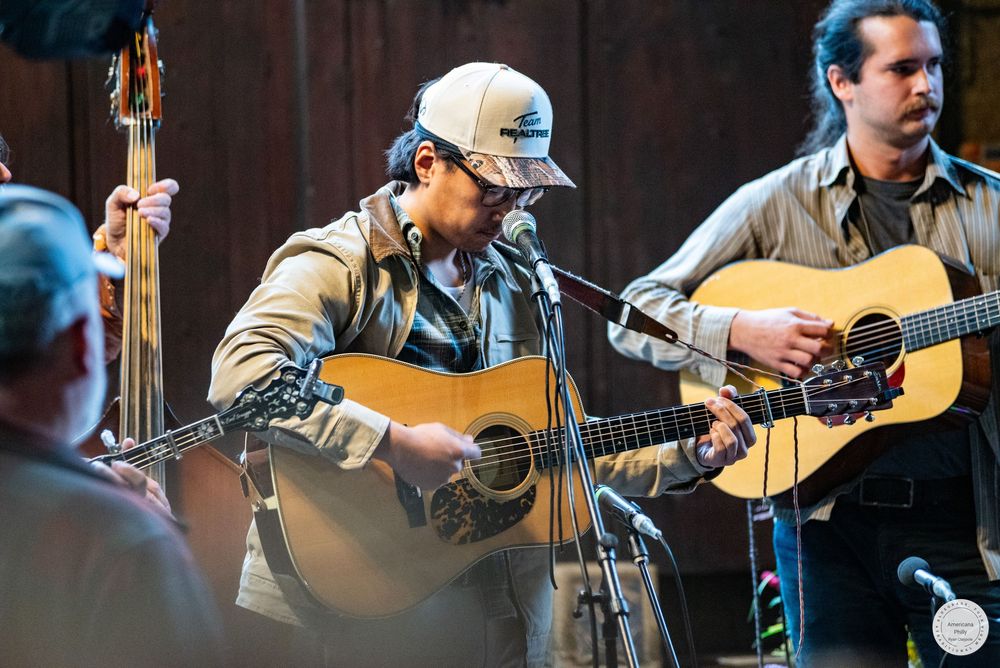 Two bluegrass musicians play acoustic guitars on stage beside a microphone, with an upright bass player visible behind them.