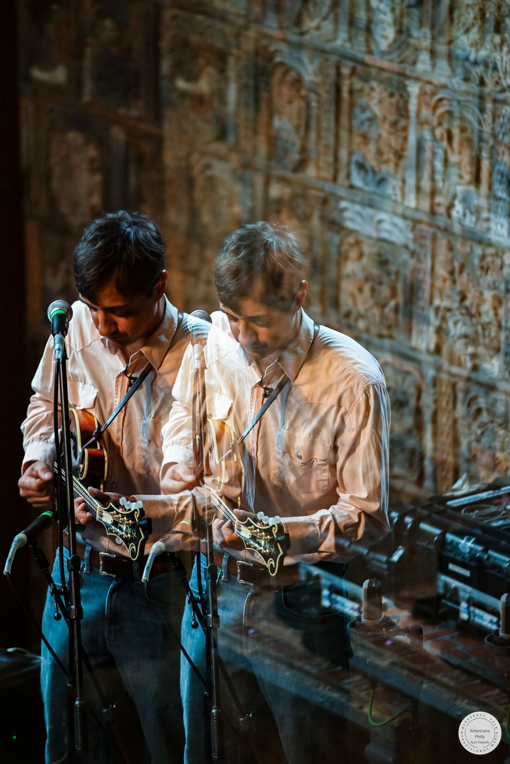 Double-exposure photo of a musician in a cream button-up shirt and jeans playing a stringed instrument beside a microphone on stage.