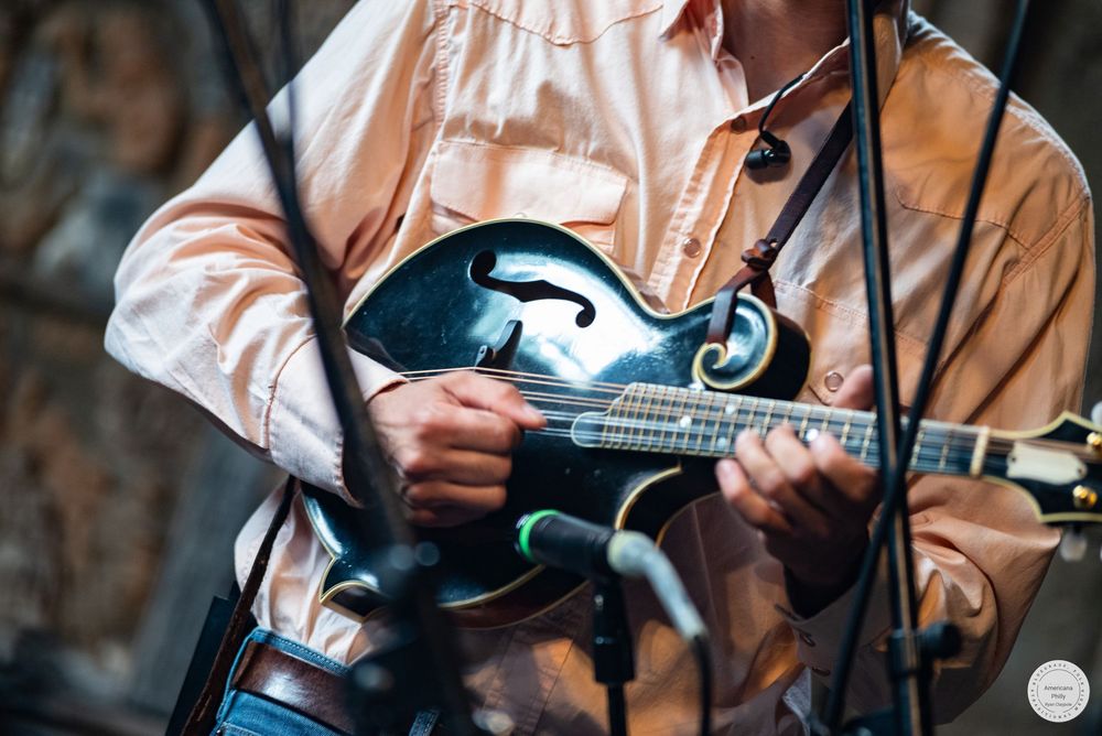 Musician in a peach button-up shirt plays a black mandolin on stage, with microphones positioned close to the instrument.