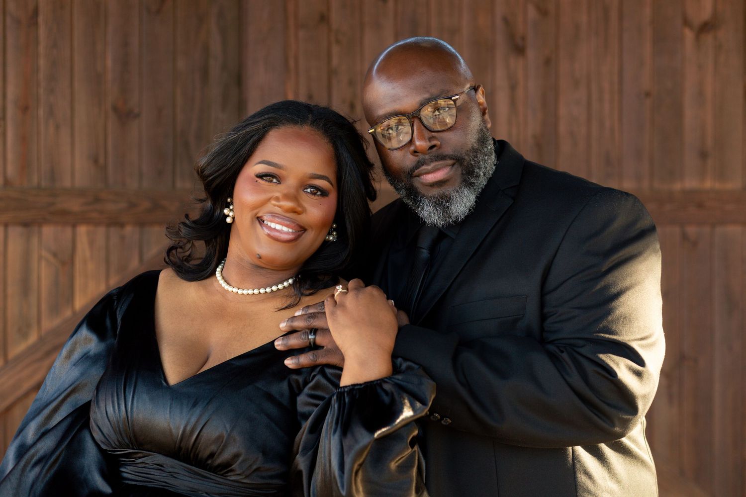 Smiling woman in a black satin dress and pearl necklace embraces a bearded man in glasses and a dark suit against a rustic wooden backdrop.