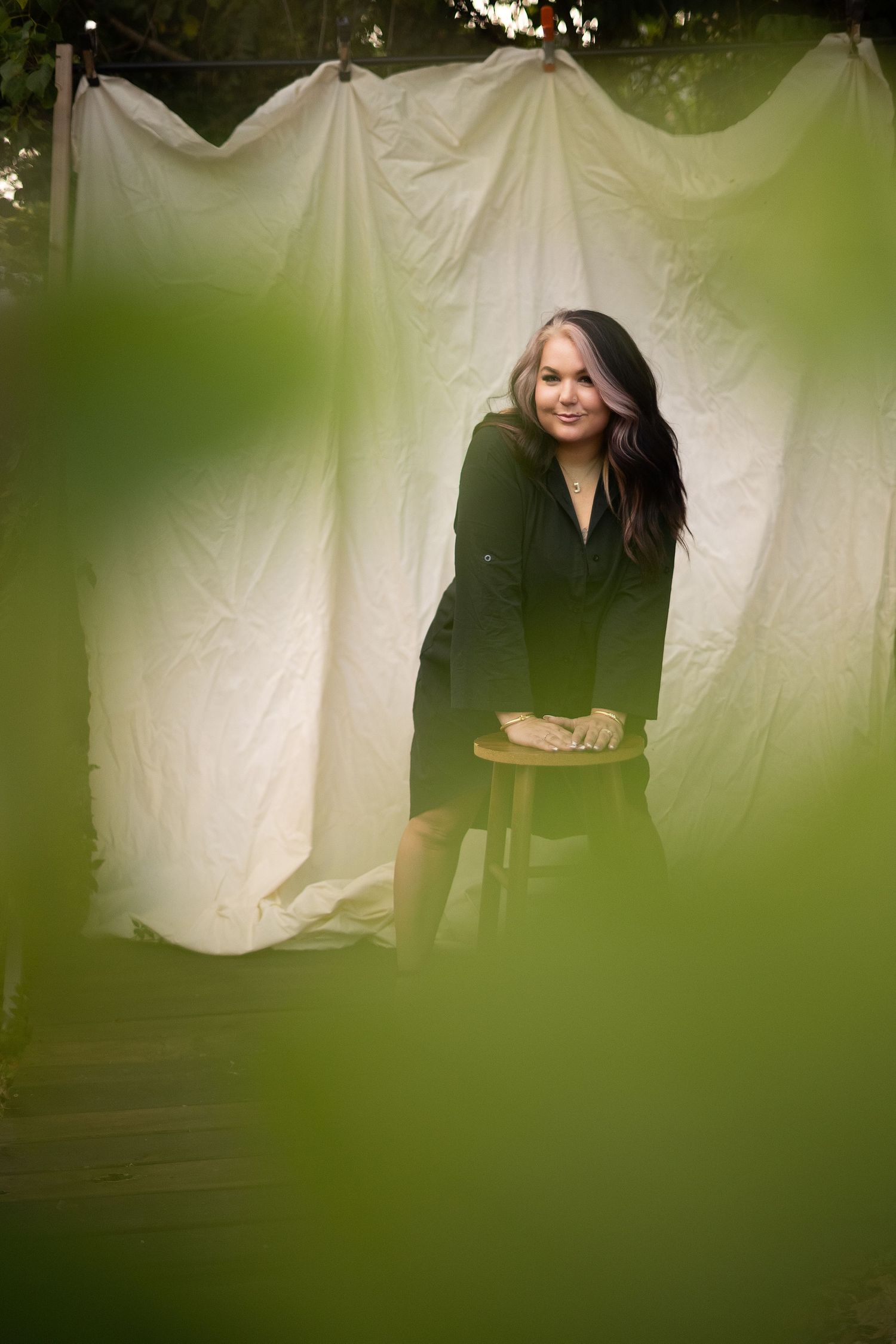 Woman in black dress leaning on wooden stool, framed by white sheets outdoors with green foliage foreground.