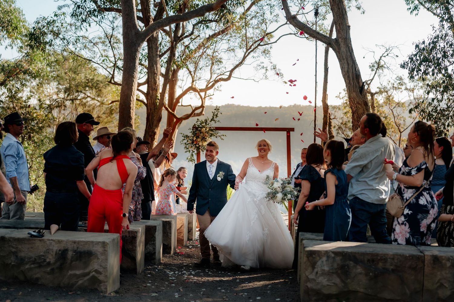 Bride and groom walk down outdoor aisle surrounded by guests throwing petals at rustic bush wedding ceremony.