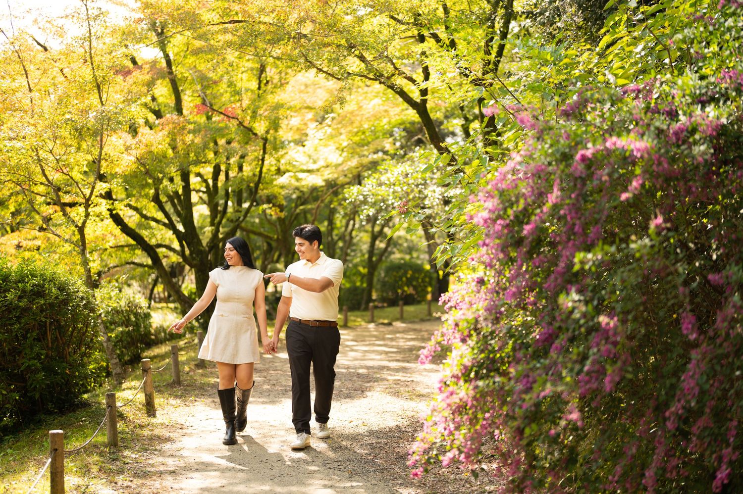 Couple holding hands and walking along a sunlit garden path lined with blooming pink flowers and golden autumn trees.