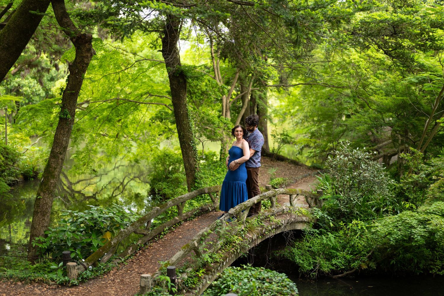 Couple in a blue dress and casual attire embrace on a stone footbridge surrounded by lush green trees in a serene garden.