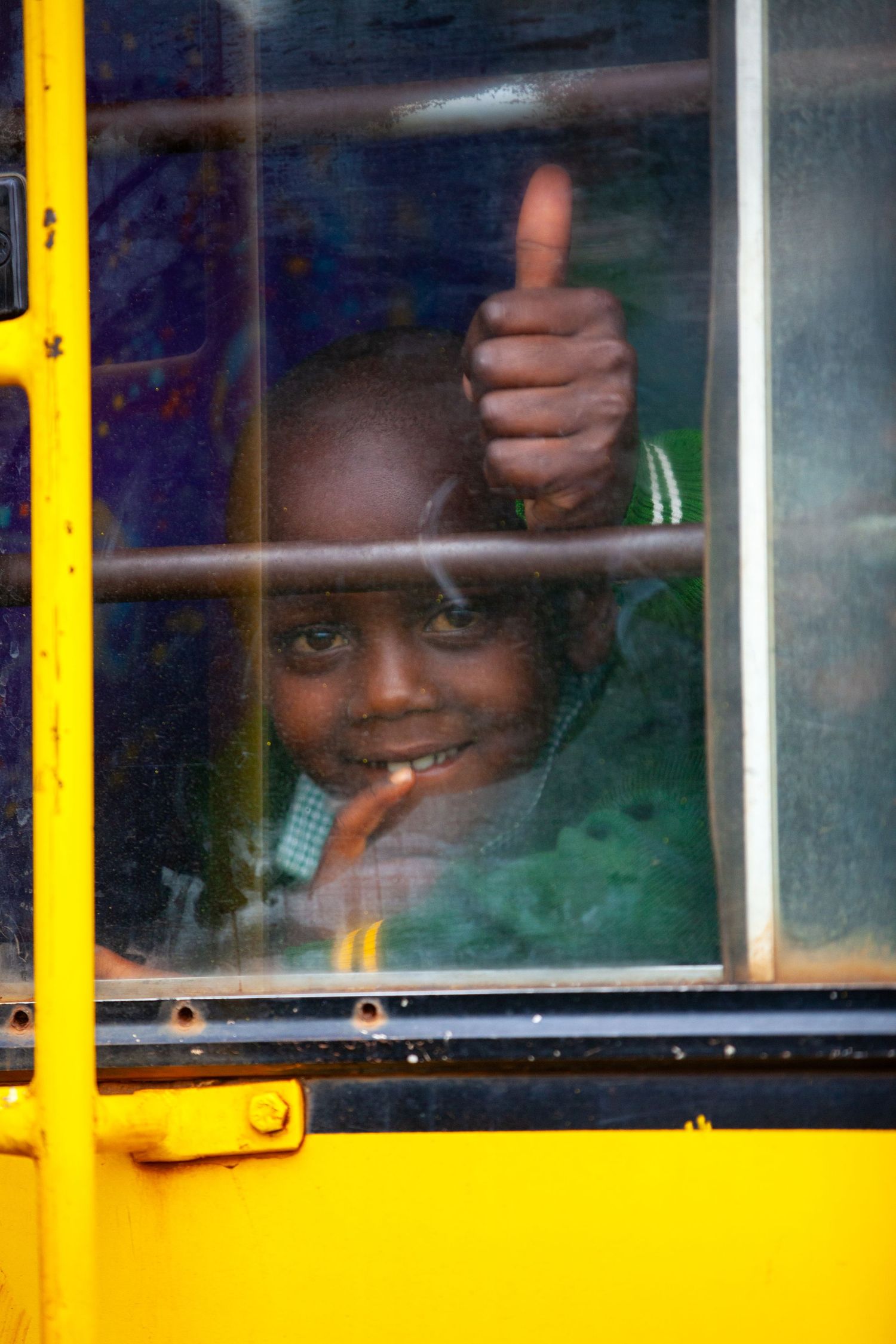 Smiling child gives a thumbs up through the window of a yellow school bus, wearing a green jacket and looking cheerfully at the camera.