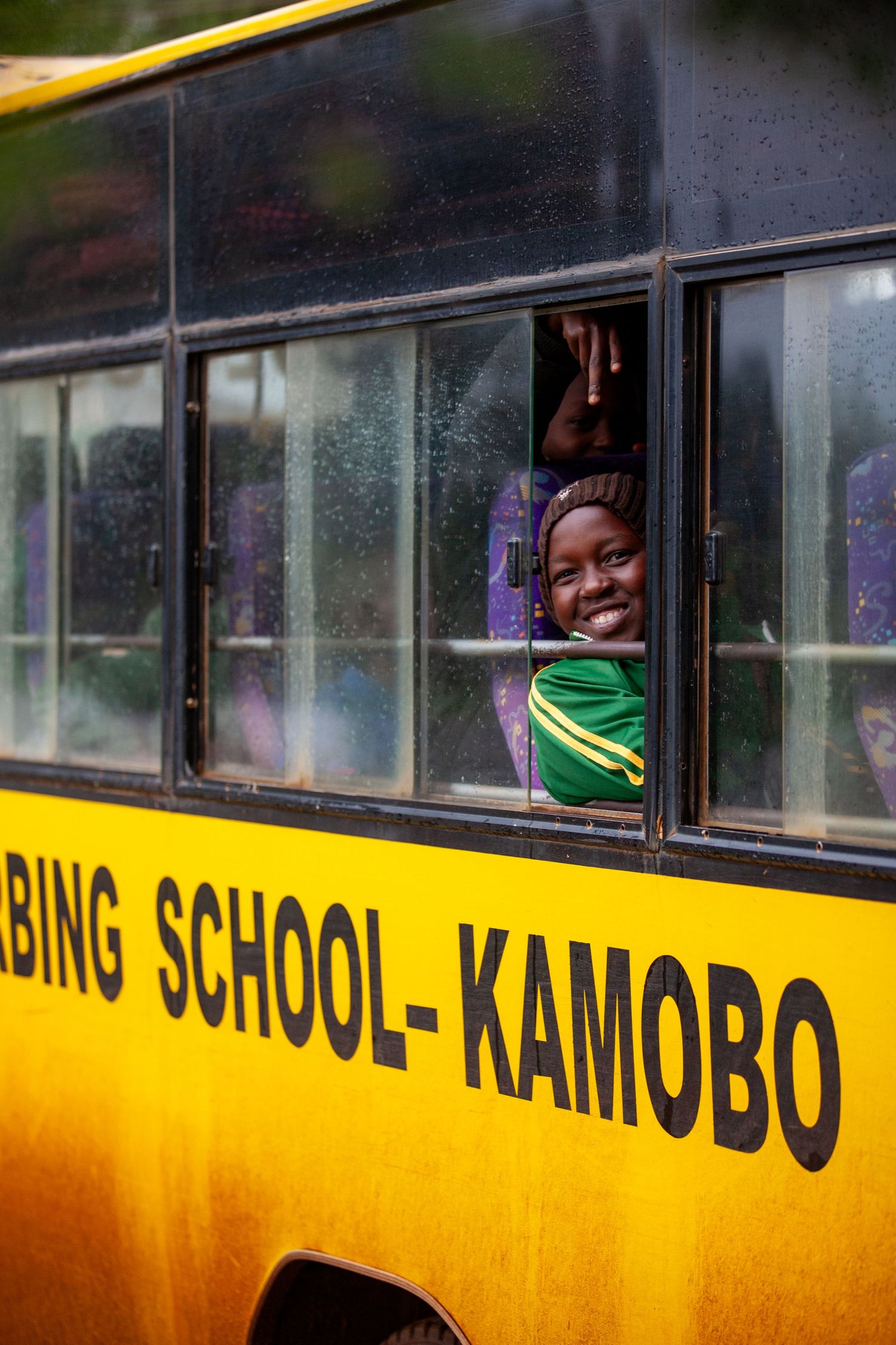 A smiling child in a green jacket peeks out of a rain-speckled window on a yellow school bus marked Kamobo.