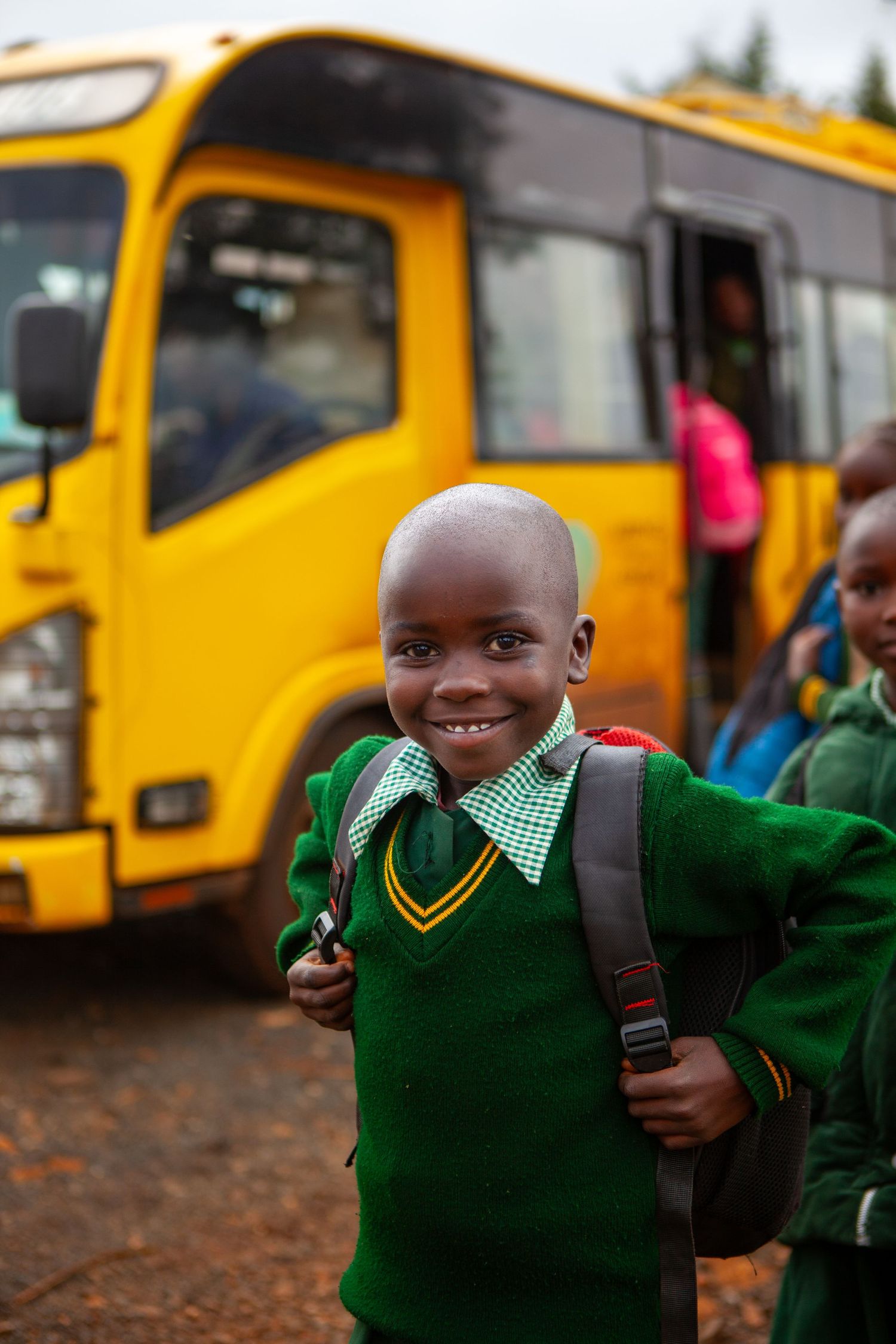 Smiling boy in a green school uniform and backpack stands in front of a yellow school bus as other students board behind him.