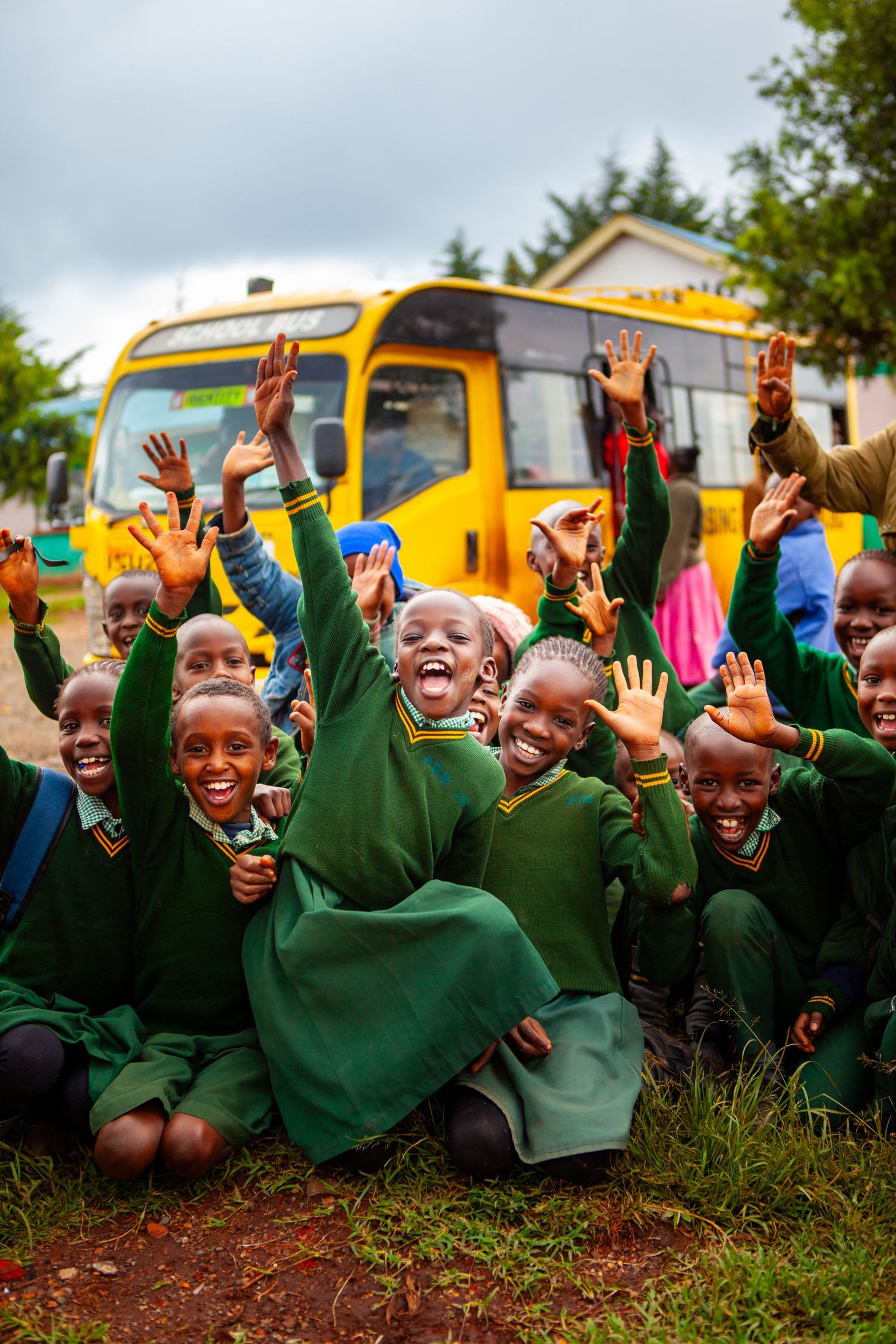Group of cheerful schoolchildren in green uniforms sit on the grass raising their hands in front of a yellow school bus.