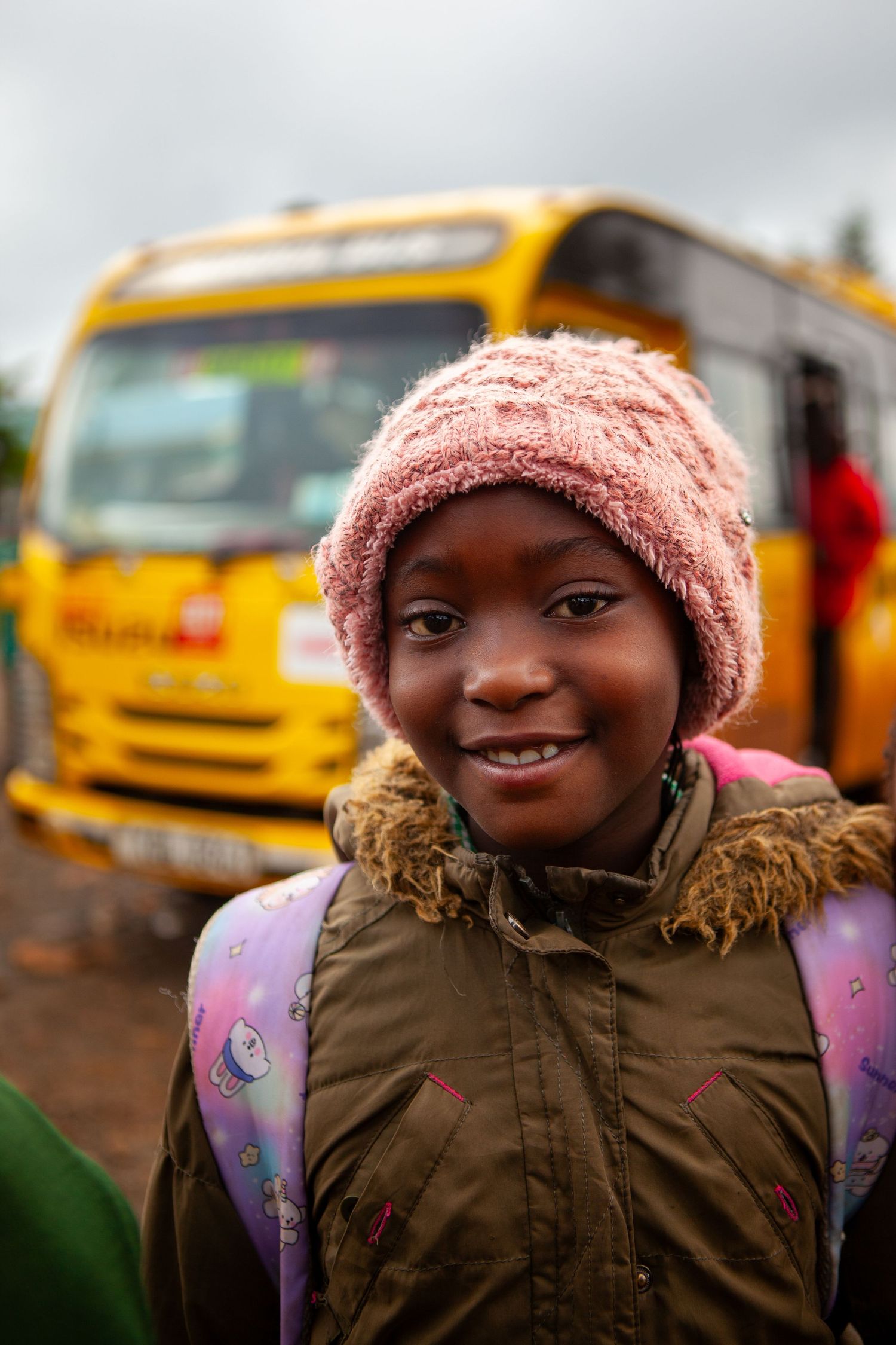 A smiling girl in a pink knit hat and olive winter coat with a backpack stands in front of a yellow school bus.