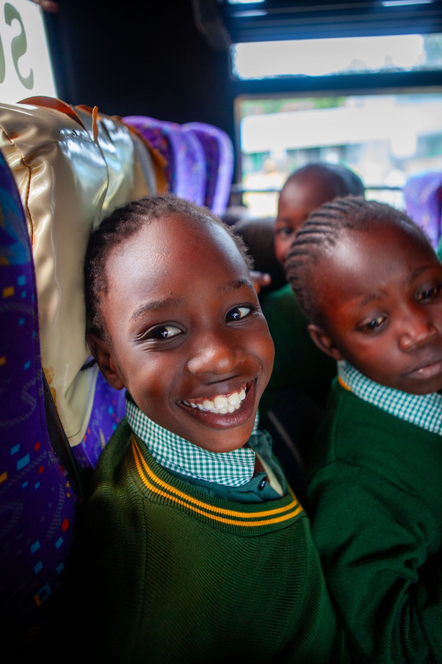 Smiling schoolgirl in a green uniform with a checkered collar sits on a bus beside classmates, beaming at the camera.
