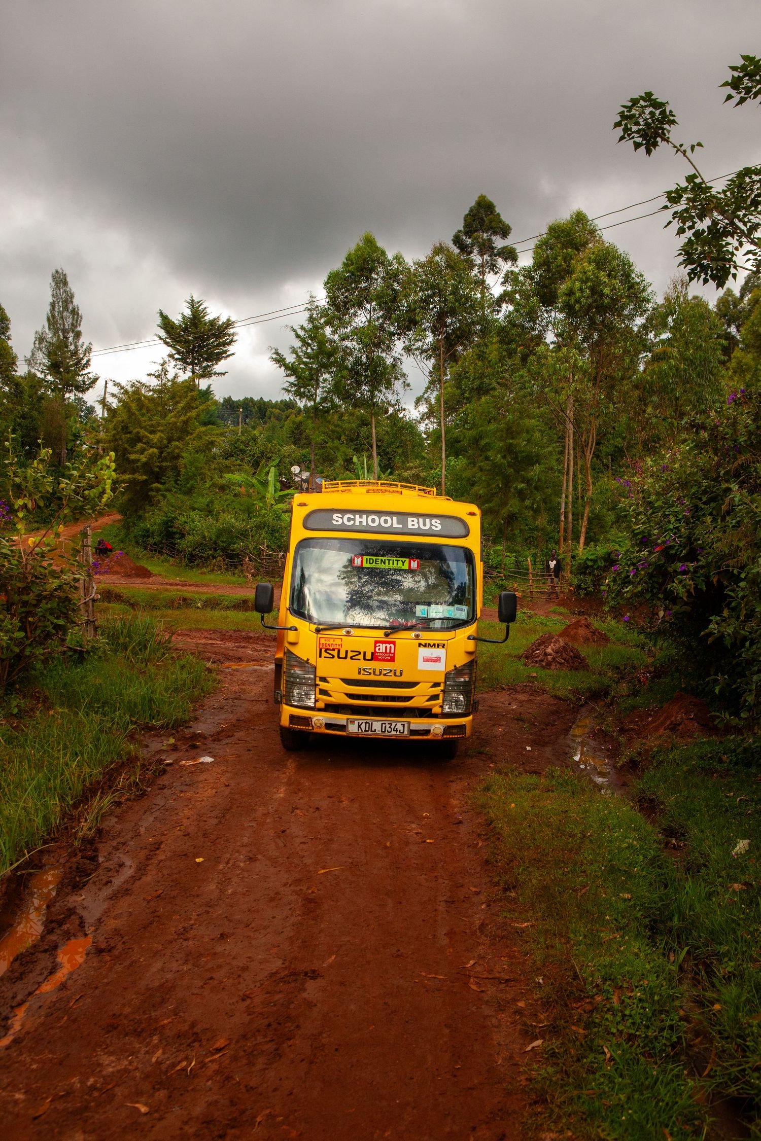 Yellow Isuzu school bus driving along a muddy red dirt road lined with green trees under a stormy, overcast sky.