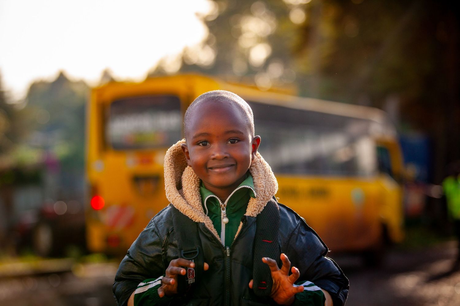Happy young child in a hooded jacket smiles outdoors with hands raised in front of a yellow school bus in warm sunlight.