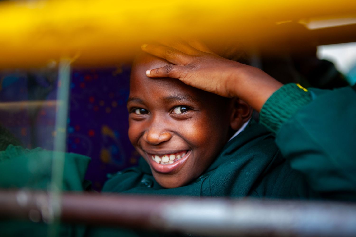 Smiling child in a green sweater peeks through yellow and metal bars, one hand resting on their forehead, with a bright, joyful expression.