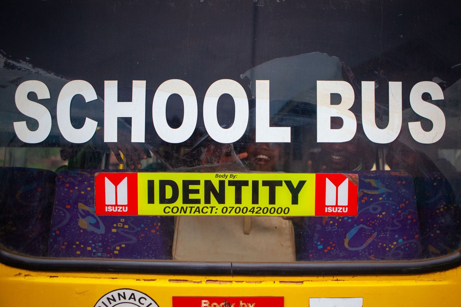 Yellow school bus rear window with white SCHOOL BUS lettering and an Isuzu Identity sticker listing a contact phone number.