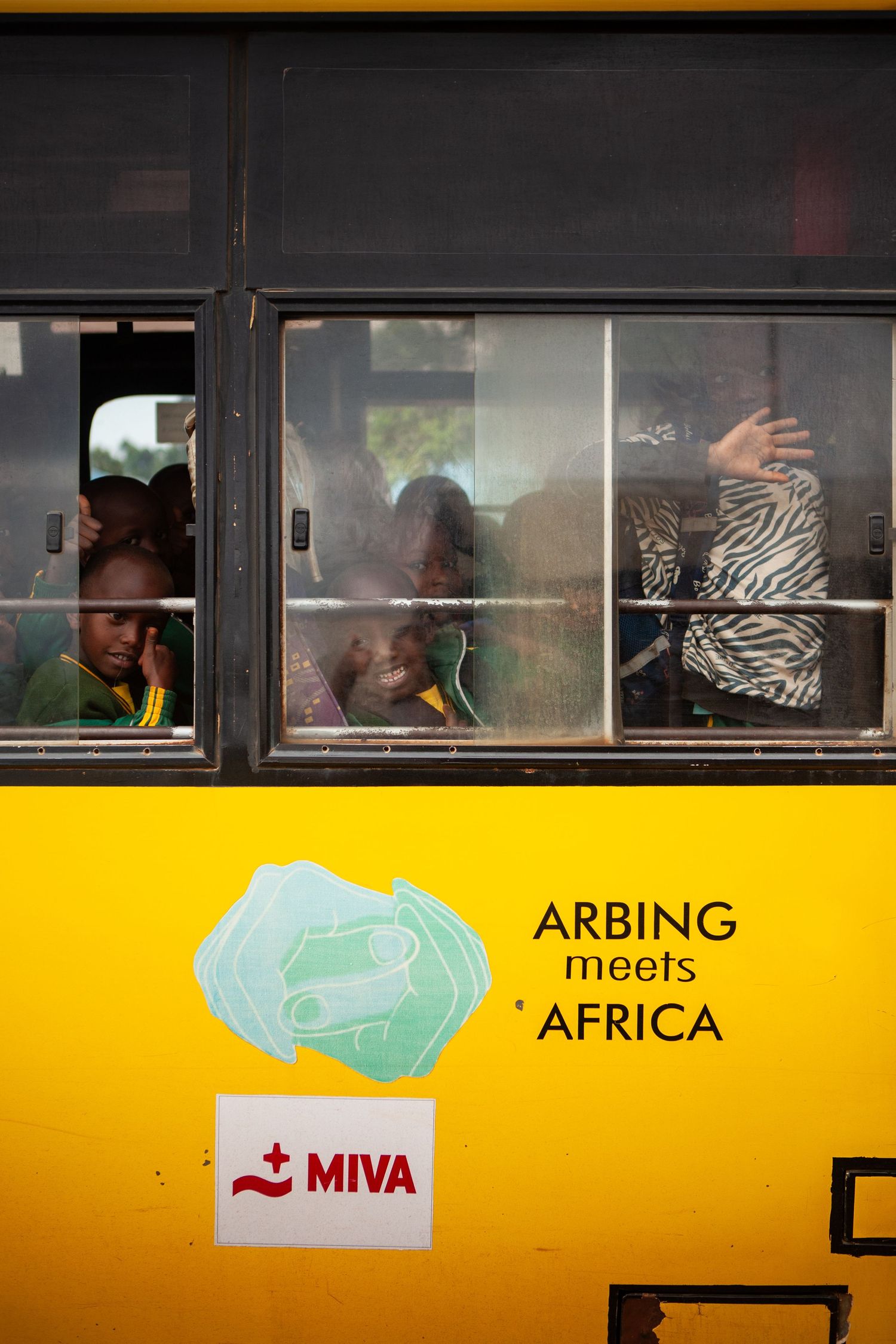 Children peer and wave through the windows of a yellow bus marked Arbing meets Africa and MIVA.