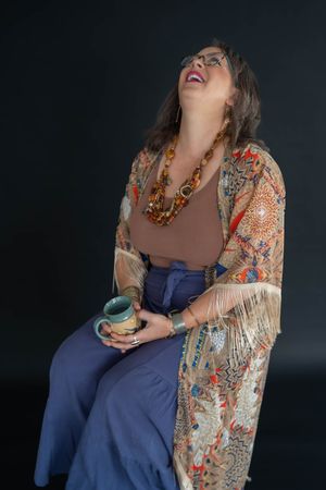 Woman laughing with head tilted back, holding a ceramic mug, wearing a fringed floral kimono, amber beaded necklace and blue wide-leg pants.