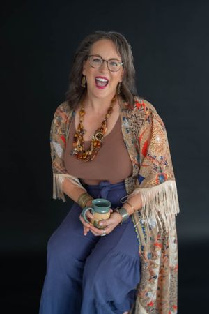 Smiling woman in glasses laughs while holding a teal ceramic mug, wearing a fringed floral kimono, beaded necklace and blue pants.