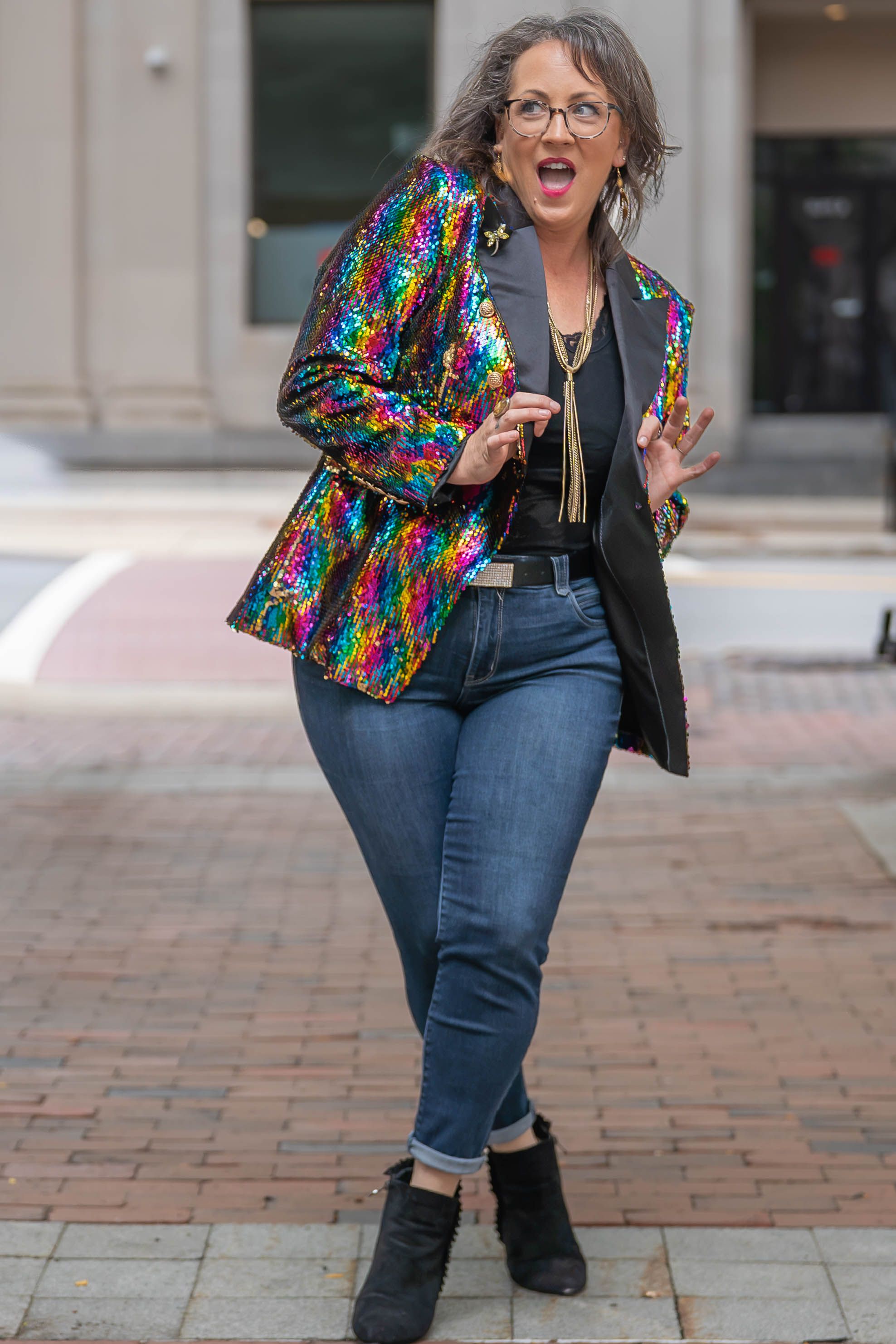Woman in glasses laughs on a brick sidewalk wearing a rainbow sequin blazer, black top, skinny jeans and black ankle boots.