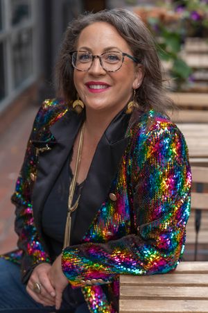 Smiling woman with glasses and gold earrings wears a sparkling rainbow sequin jacket while sitting at an outdoor wooden table.