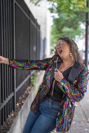 Woman in a rainbow sequin blazer and glasses leans against a black iron fence on a tree-lined city sidewalk, singing with joy.