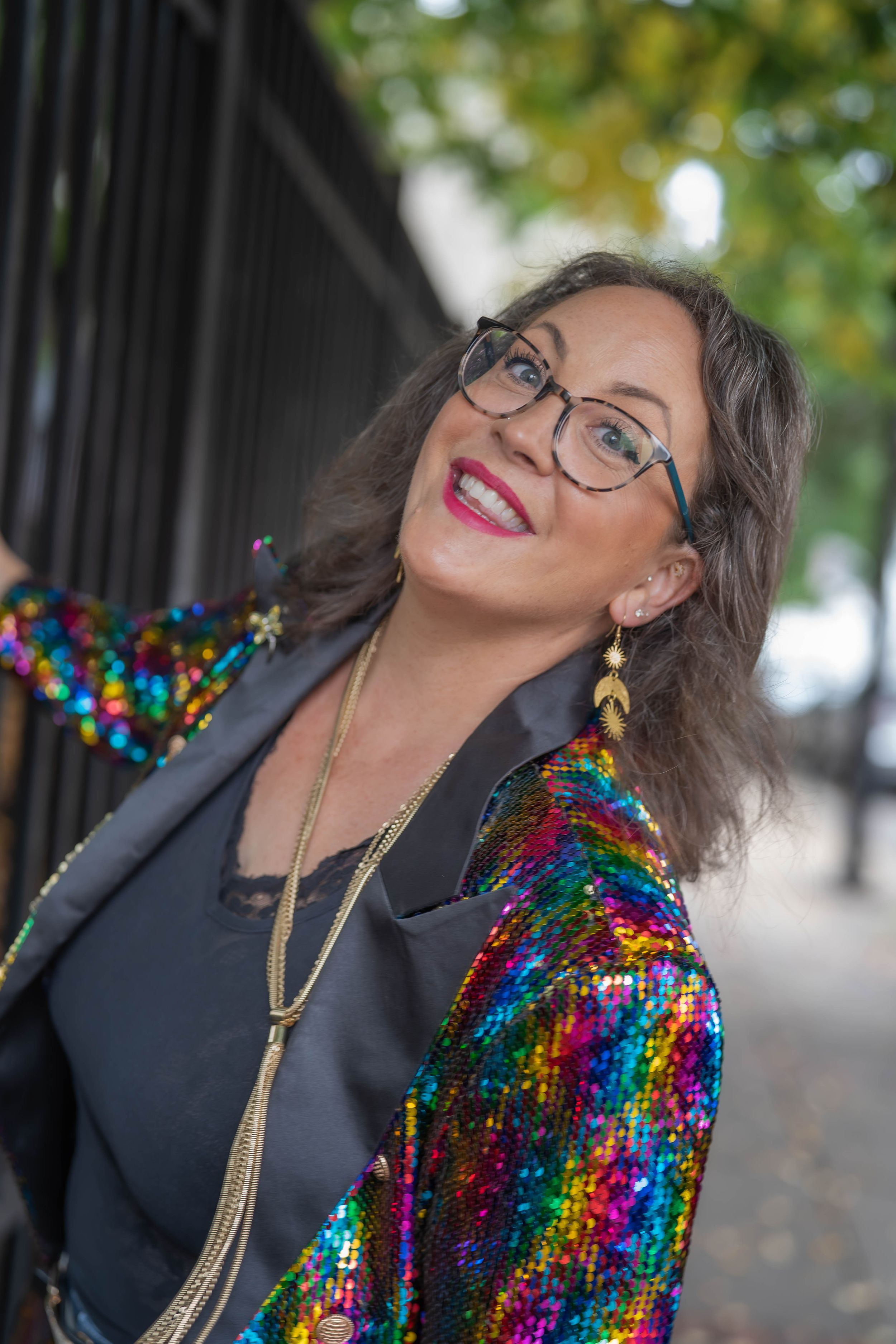 Smiling woman with glasses and red lipstick wears a rainbow sequin jacket, gold tassel necklace and leaf earrings beside a black iron fence.
