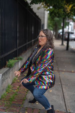 Woman in glasses and a rainbow sequin blazer squats playfully on a brick sidewalk beside an iron fence on a tree-lined city street.