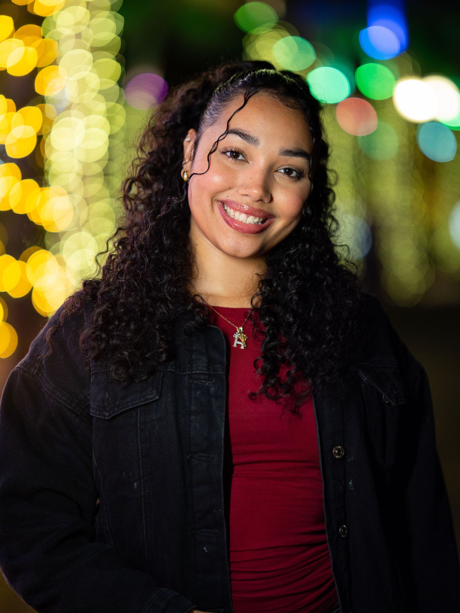 Smiling young woman with long curly dark hair, a denim jacket, and a red top stands before glowing bokeh string lights at night.