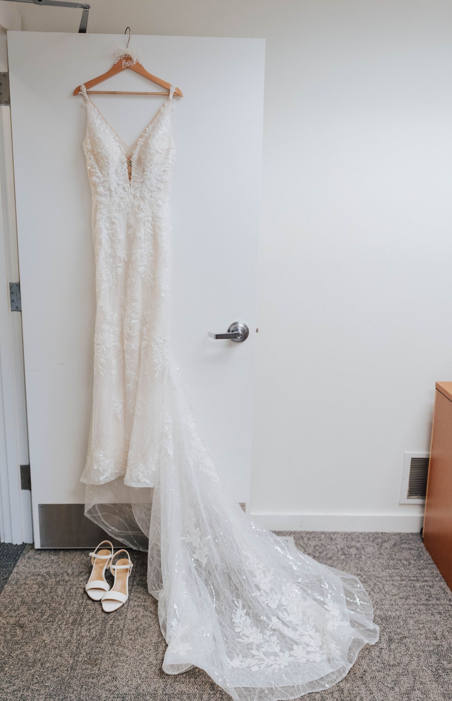 White lace wedding gown with train hanging on door, white bridal heels on floor below.